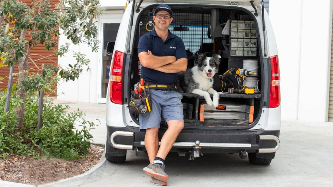 A tradesperson leaning against the back of an open van with tools and equipment visible inside, alongside a dog sitting inside the van.