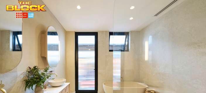 A modern bathroom with a bath with a table next to it, windows, two mirrors on the wall, two sinks with drawers underneath them in it.