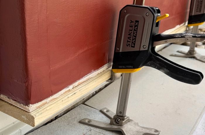 Timber trim clamped to the base of a waterproofed red panel near a white wall.