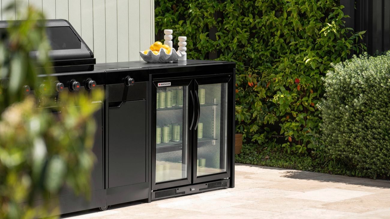 A outdoor kitchen with a barbecue and bar fridge in it sitting on a paved area, with plants around the paving.
