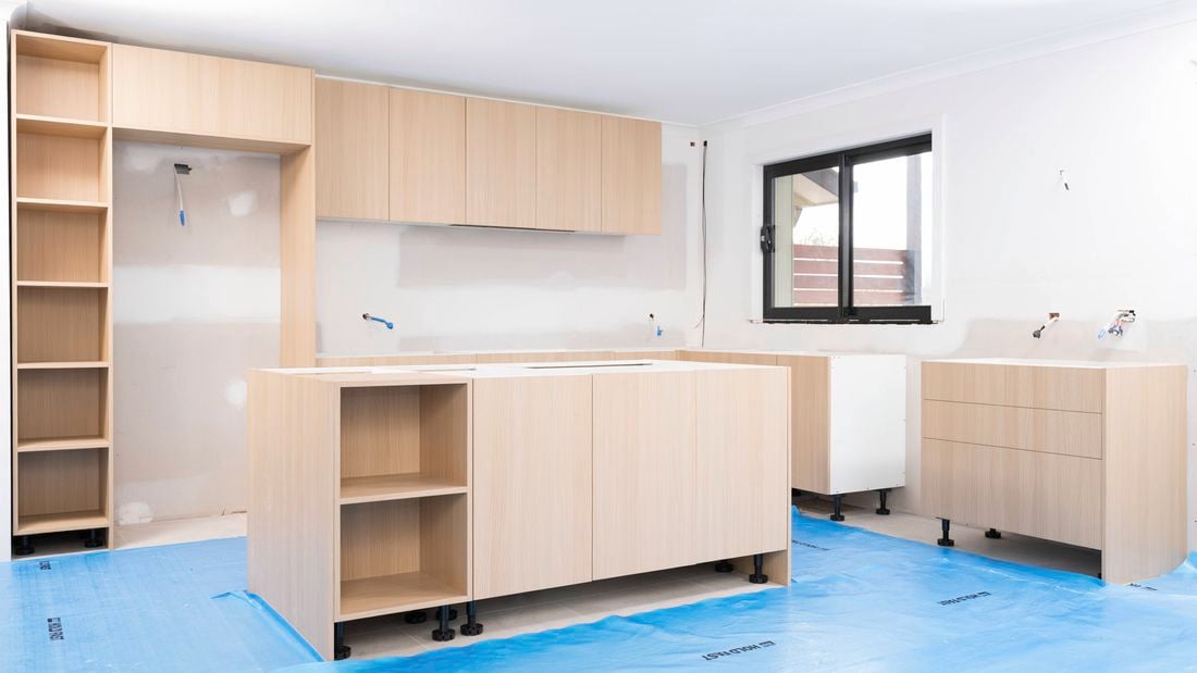 The image shows a kitchen under renovation, with new light wood cabinetry being installed and blue protective sheets covering the floor