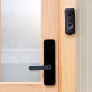 A black Swann video doorbell is mounted beside the handle of a wooden front door with frosted glass panels. 