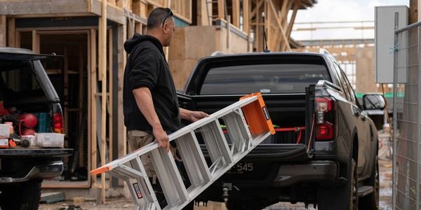 A tradie in work boots is putting a Citeco ladder into the back of a work ute on a job site.
