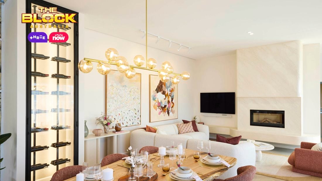 A modern living and dining area featuring a dining table set with plates, glasses, and candles, surrounded by six chairs. A contemporary chandelier with spherical lights hangs above. In the background, a white sofa with colorful cushions sits beneath two large abstract artworks. A fireplace with a TV above it is on the right, and a tall wine rack filled with bottles is on the left.