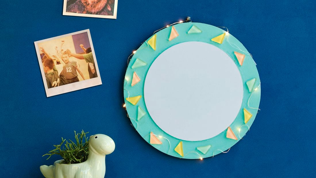 A round decorative wall mirror with a teal painted frame, adorned with pastel triangle embellishments and string lights, hanging on a blue wall next to two polaroid photos and a dinosaur-shaped plant pot