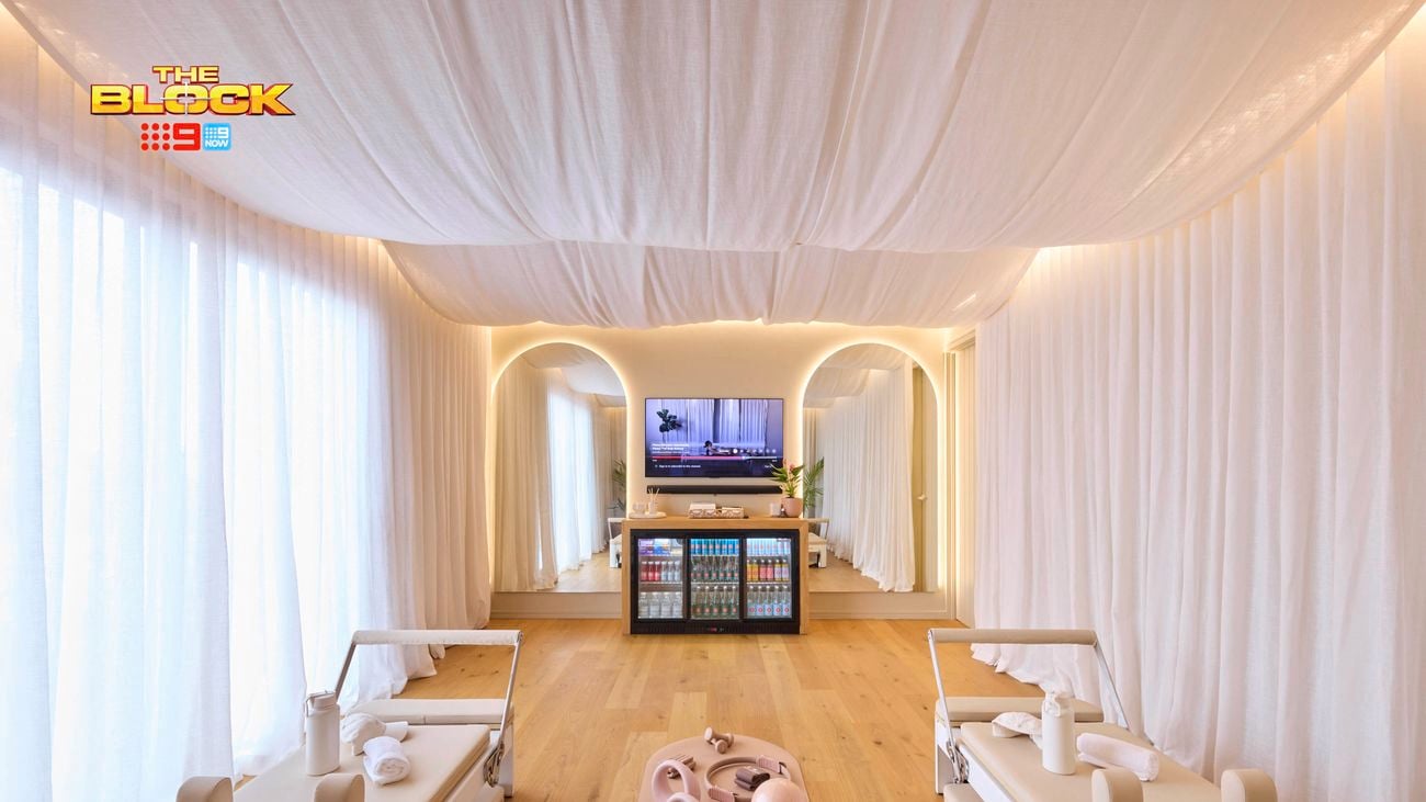 A modern Pilates studio with two reformer machines flanking a central yoga mat. The space features light wooden flooring, white curtains, and a ceiling draped with white fabric for a calming ambiance. At the far end, a mirrored wall with an arched design sits below a mounted TV and a cabinet stocked with beverages. 