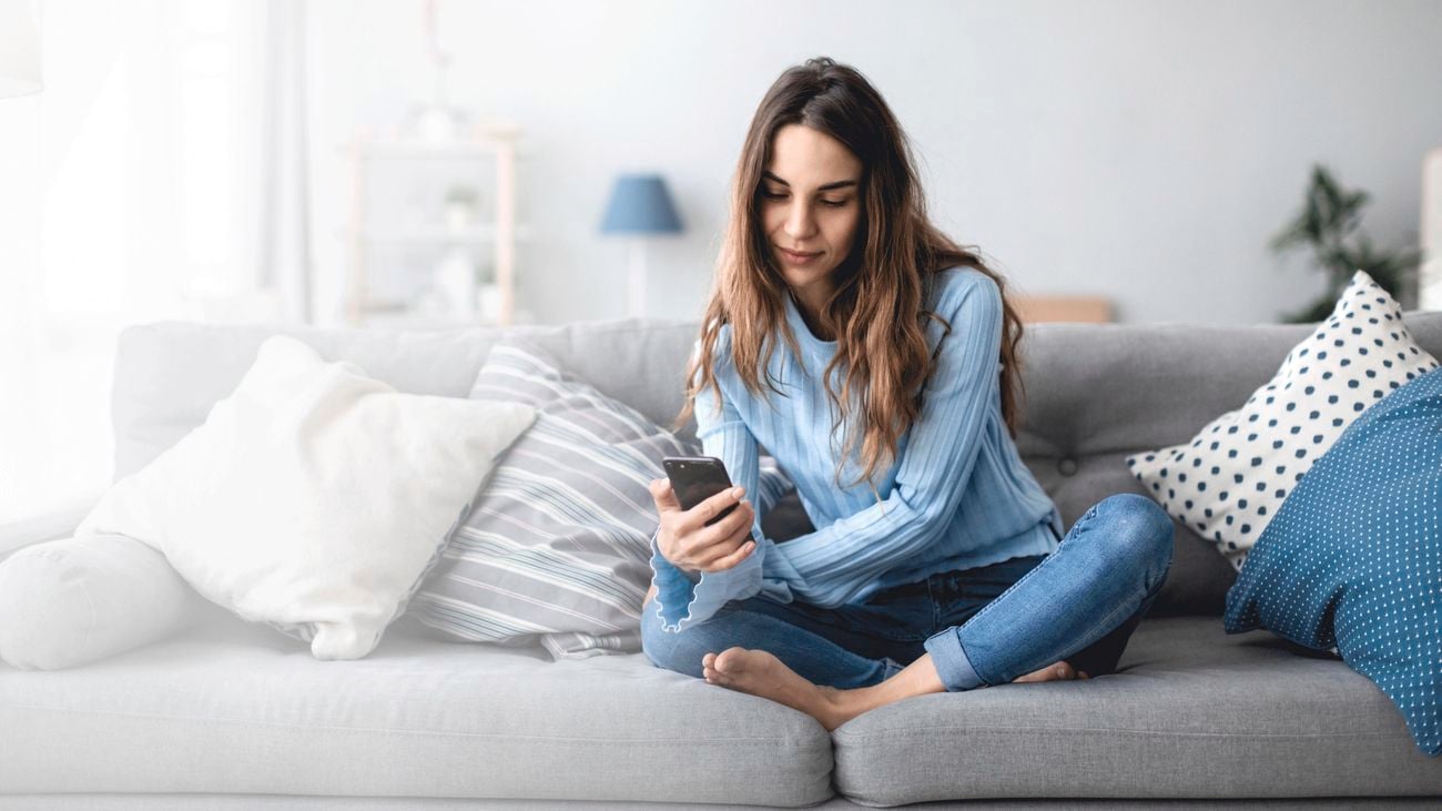 Person sitting cross-legged on a grey couch with cushions, holding a smartphone