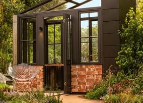 Modern black-framed greenhouse garden room with brick base, surrounded by lush plantings.