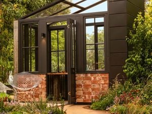 Modern black-framed greenhouse garden room with brick base, surrounded by lush plantings.