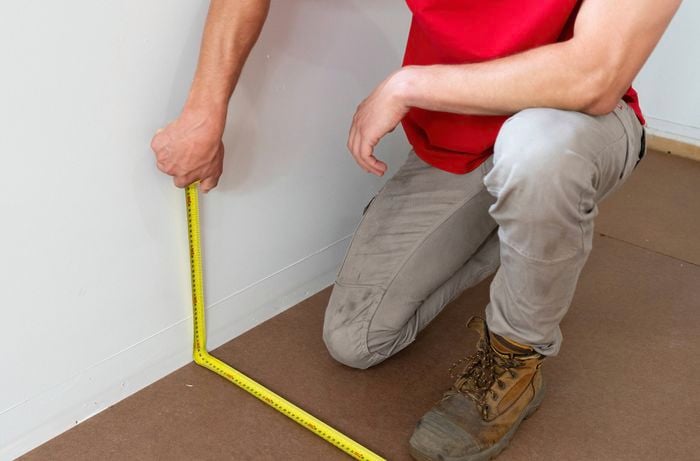 A person in a red shirt and work boots is measuring the length and width of the room with tape measure.