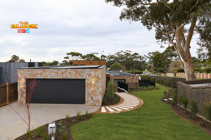 Modern garage with natural stone cladding, a black roller door, curved stepping stone pathway, landscaped lawn and mature trees.