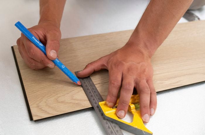 A person is marking a cut line on a laminate floorboard using a combination square and a blue pencil. 