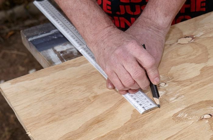A person marking a sheet of plywood using a ruler and pencil