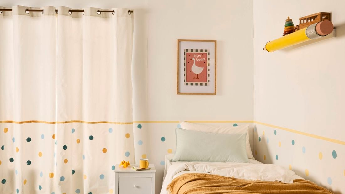 A large yellow pencil shelf topped with wooden toys on a neutral wall with colourful polka dots with a bed with pillows and a cover on it, in the bedroom.