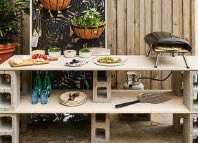 A besser block outdoor kitchen bench space with pizza oven, utensils, bowls of herbs and a timber chopping board with vegetables on it.