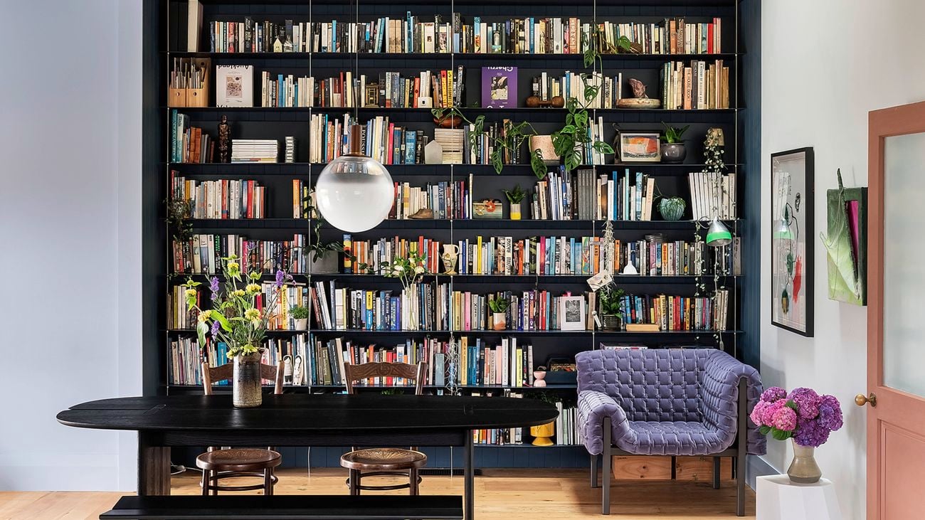 Room with a floor-to-ceiling bookcase, a few plants and a globe pendant. Long black table with two chairs on a patterned rug, purple armchair to the right, vases of flowers, pale walls, and a timber floor