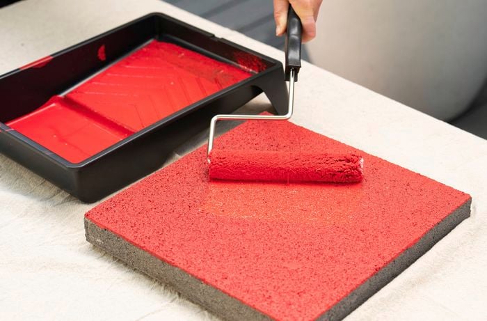 A person is painting a concrete paver with bright red colour using a paint roller. 