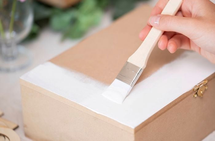 A person is applying a white base coat to a wooden craft box using a paint brush as part of creating a customised DIY memory box.