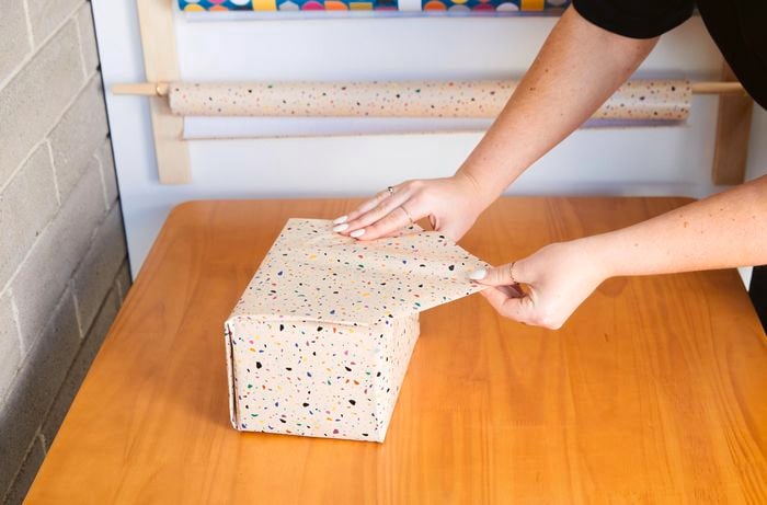 One hand stabilises a box wrapped in wrapping paper, while the other hand pulls a folded edge of wrapping paper over the side of the box.