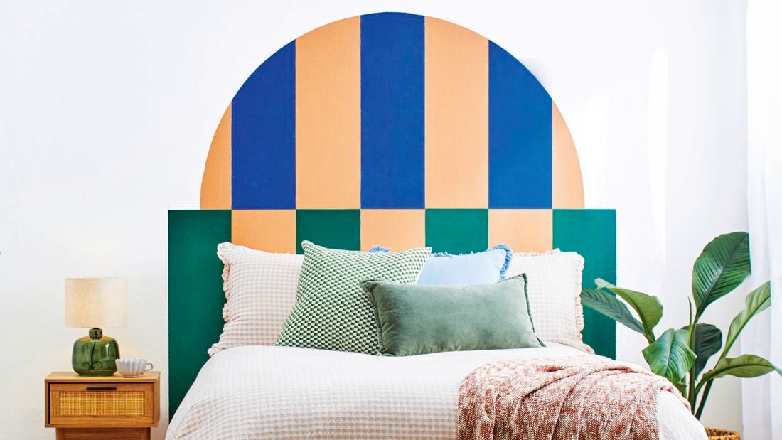 Bedroom with a painted geometric half-arch headboard in blue, peach and green, white bedding with layered cushions, bedside table and indoor plant.