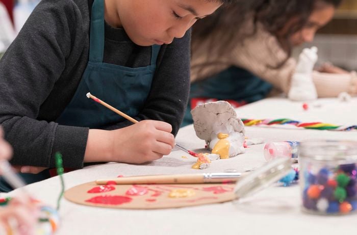 A child wearing a dark shirt and apron focuses on painting a clay creature at a table filled with colorful craft supplies.