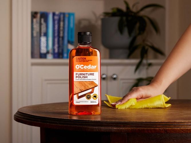 A person's hand holding a cloth against a wooden table with a bottle of O'Cedar furniture polish on it.