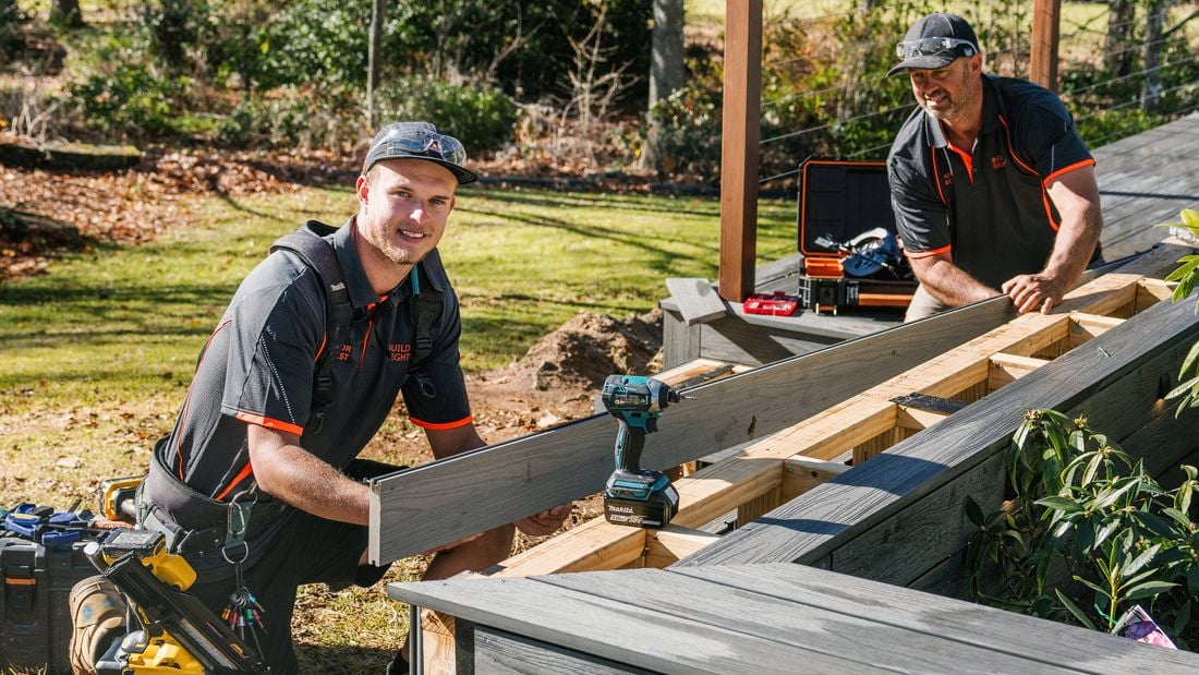 Two tradespeople install grey composite decking on a timber frame in a backyard, one holding a board while the other works nearby. Power tools and a tool case sit on the structure, with grass and trees behind.