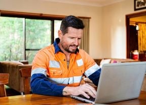 A man in hi-vis working on a laptop placed on a dining table inside a home