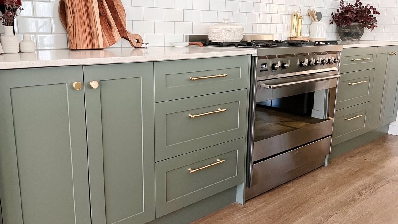 A kitchen with white tiles on the wall, green cupboards and drawers and timber flooring.