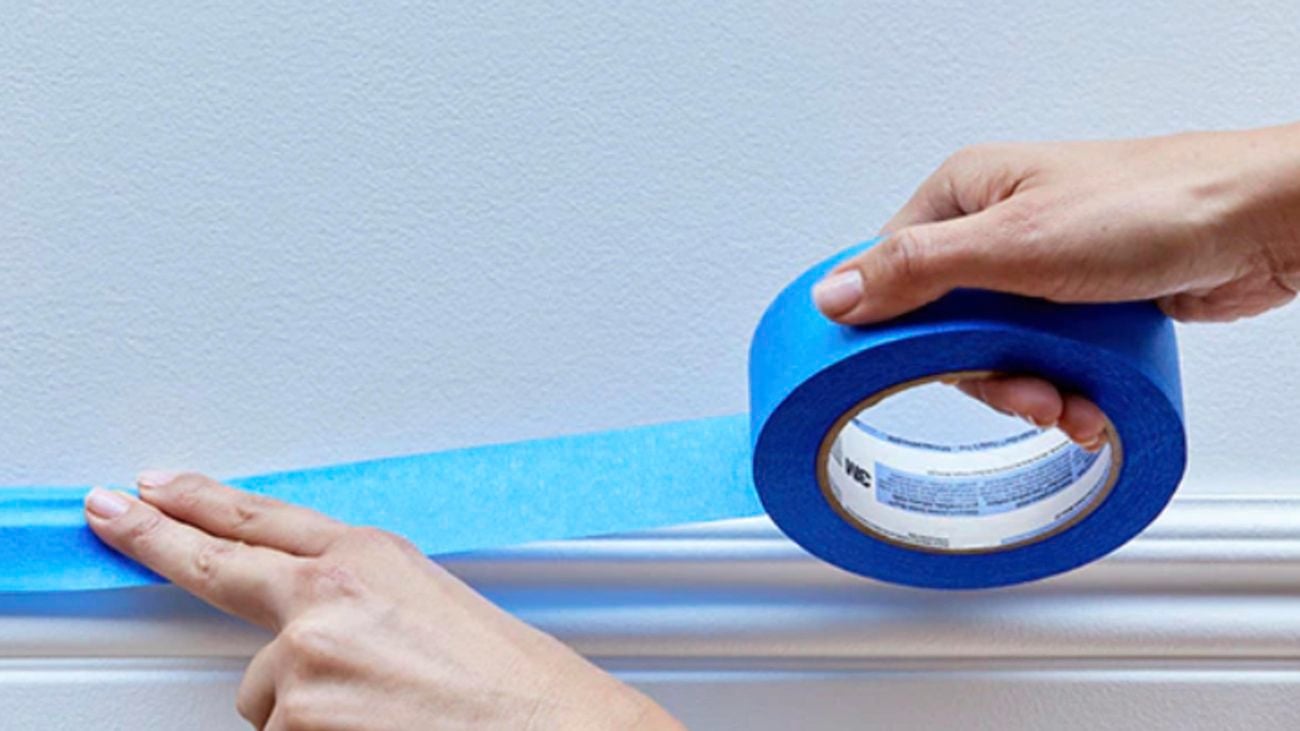 A hand applying blue Scotch tape along the top edge of a skirting board