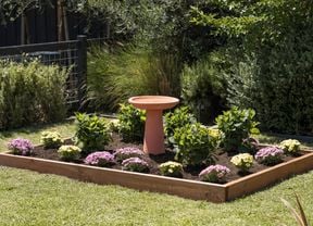 A raised garden bed featuring pink and yellow flowers, green shrubs, and a terracotta birdbath.