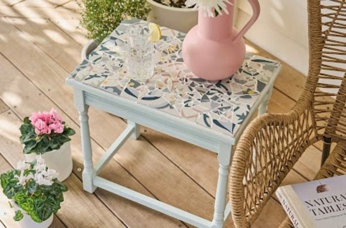 A D.I.Y mosaic tiled table sitting on a wooden deck next to a cane chair.