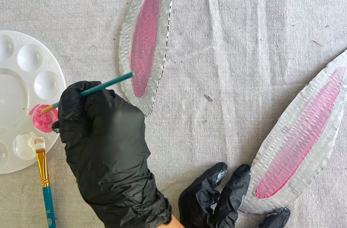 Black gloved hands apply pink paint to white bunny ears made from cardboard.