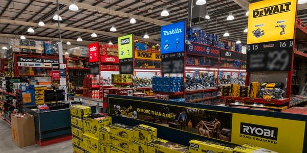 Interior of a Bunnings Warehouse power tools department featuring branded displays for Ryobi, Makita, DeWalt, Bosch and Ozito. Stacks of boxed power tools are displayed in the foreground, with overhead aisle signage and warehouse-style shelving stocked with tools and accessories throughout the store.
