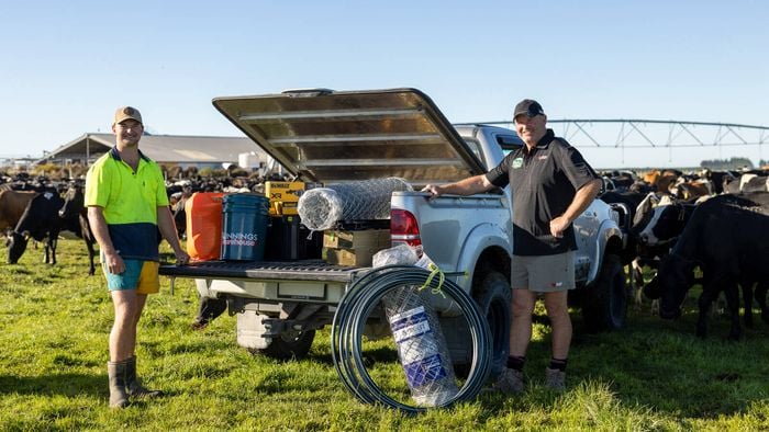 Two men standing beside an open ute on a farm, with cattle in the background; the vehicle is loaded with supplies including fencing wire, buckets, and tools.