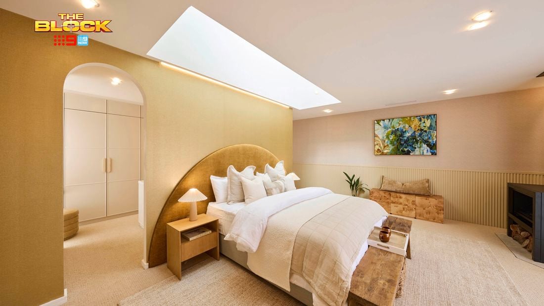 A tastefully decorated bedroom with a large bed centered under a skylight, dressed in a beige blanket and multiple pillows. Two bedside tables with lamps flank the bed. A fireplace is visible on the right, with a bench at the foot of the bed. A colorful painting hangs above a small plant and a piece of furniture. The room features neutral-toned walls and carpeted flooring.