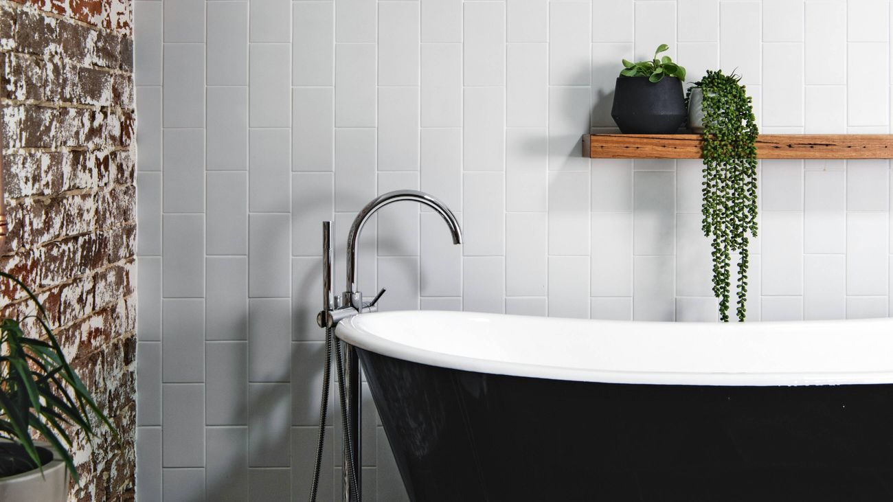 A bathroom featuring a black freestanding bathtub with a chrome tap and showerhead. Exposed brick and white tile walls in the background, with a wooden shelf with potted plants mounted above the tub.
