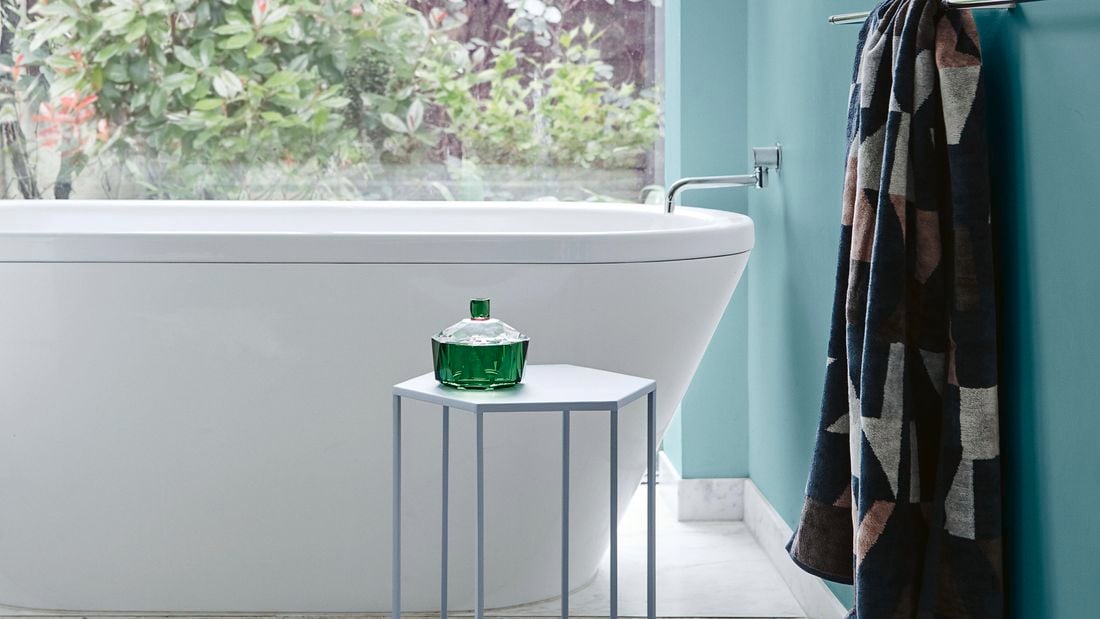 A bathroom with soft green feature wall, white bath, towel rail and small tool next to bath.
