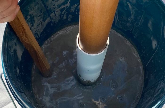 A Bunnings 20L bucket with a white pipe in the centre and a hand stirring concrete.