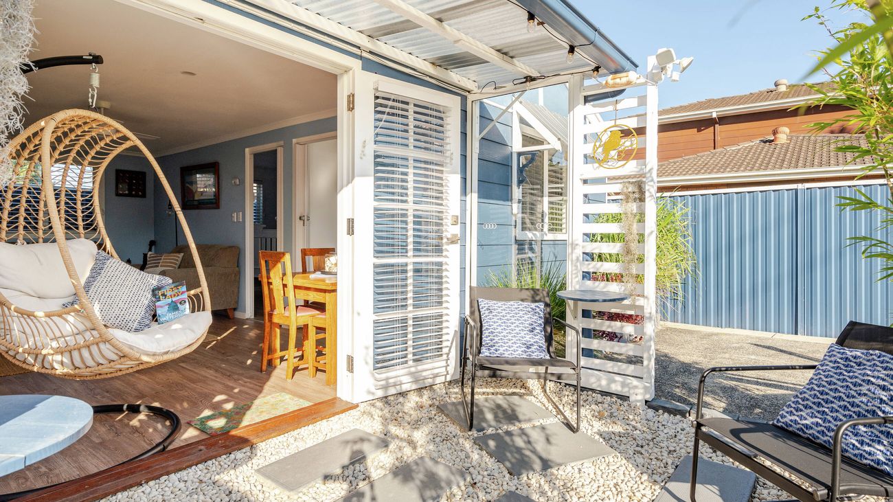 Exterior view of an outdoor patio area with a hanging wicker chair, stone pavers, outdoor seating, and an open doorway leading inside.