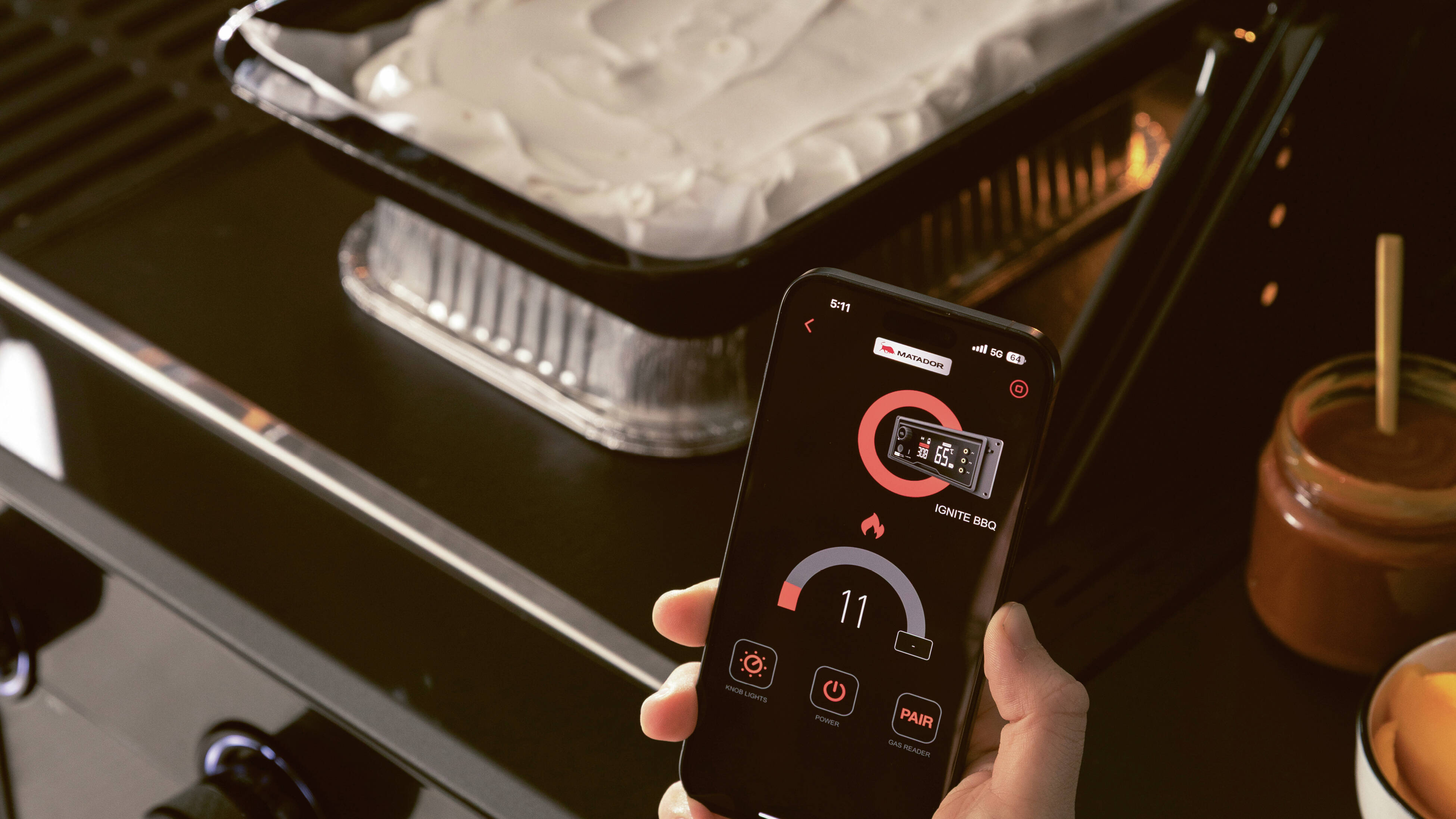 Hand holding a smartphone controlling a barbecue with food on the grill