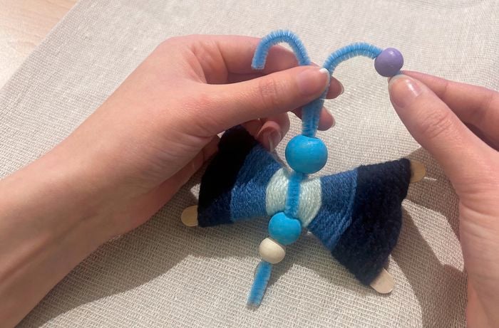 Two hands hold two blue pipe cleaners in place, with a purple bead on the right pipe cleaner.
