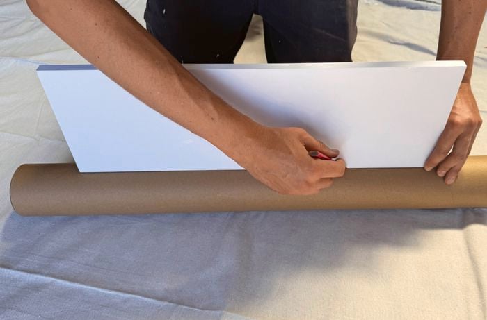 Hands holding a white shelf on a cardboard packing tube and using a pencil to mark its outline.