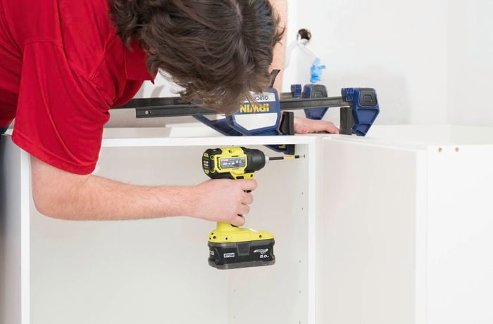 A person in a red t-shirt is using a power drill to join the two white cabinets together.