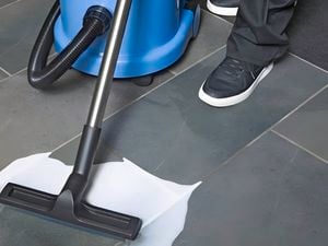 Person using a blue and black Numatic NX vacuum cleaner to clean a liquid spill on a tiled floor.