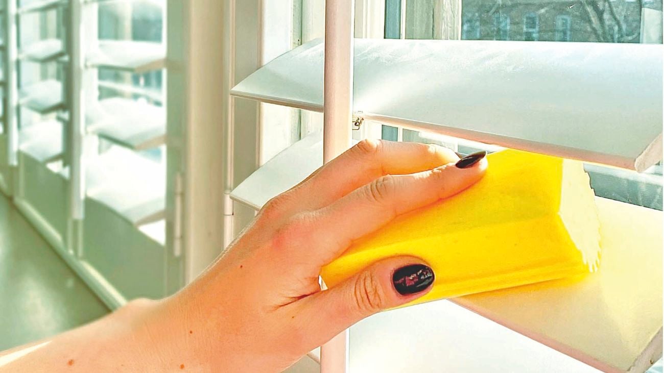 Hand uses yellow sponge to wipe dust off blinds.