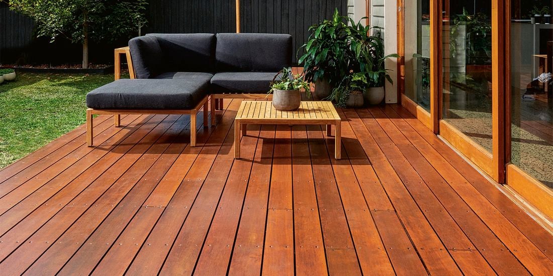 A stained backyard deck complete with timber outdoor coffee table, outdoor sofas and pot plants, with a lawn and tree in the background