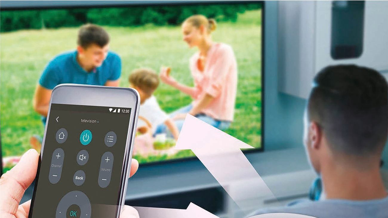 Hand holds phone with circular, smart-controller sitting on wooden bench with TV in background