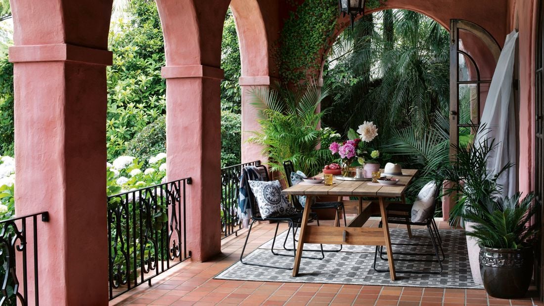 A table with crockery and a vase with flowers on it and chairs on a verandah at the back of a house.