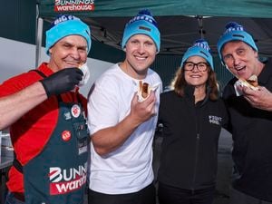 Four people wearing Biz Freeze blue beanies standing together under a Bunnings marquee, smiling while holding sausages in bread outdoors.